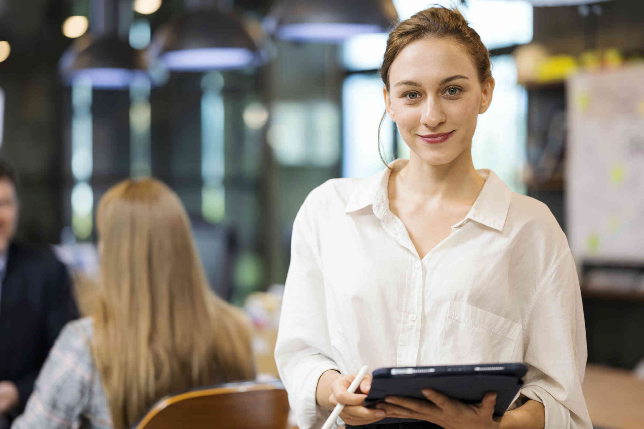 A woman in a buttondown shirt stands in an office space while holding a tablet and smiling softly at the camera.