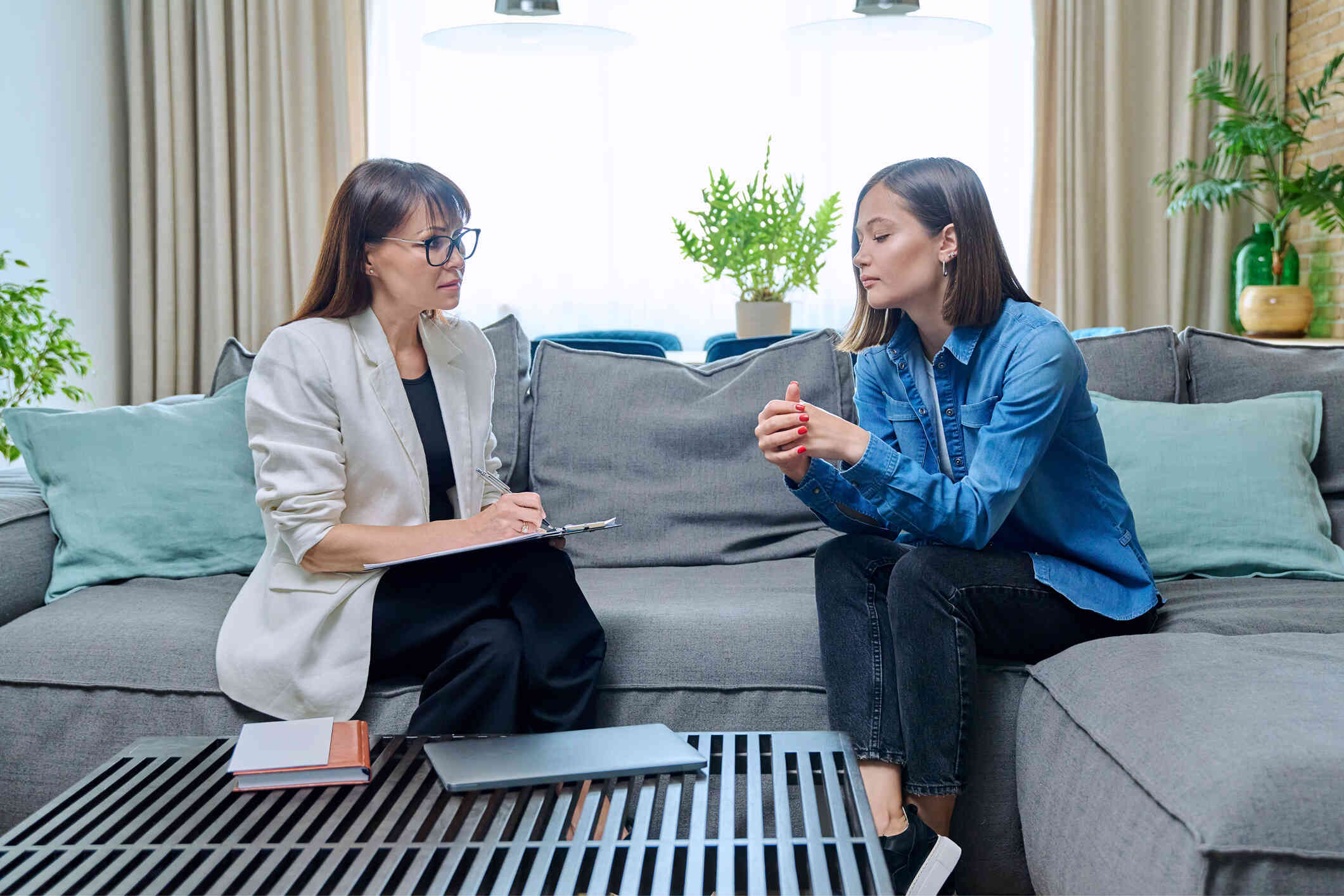 A woman ina jean shirt sits next to her female therapist on a couch while talking during a therapy session.